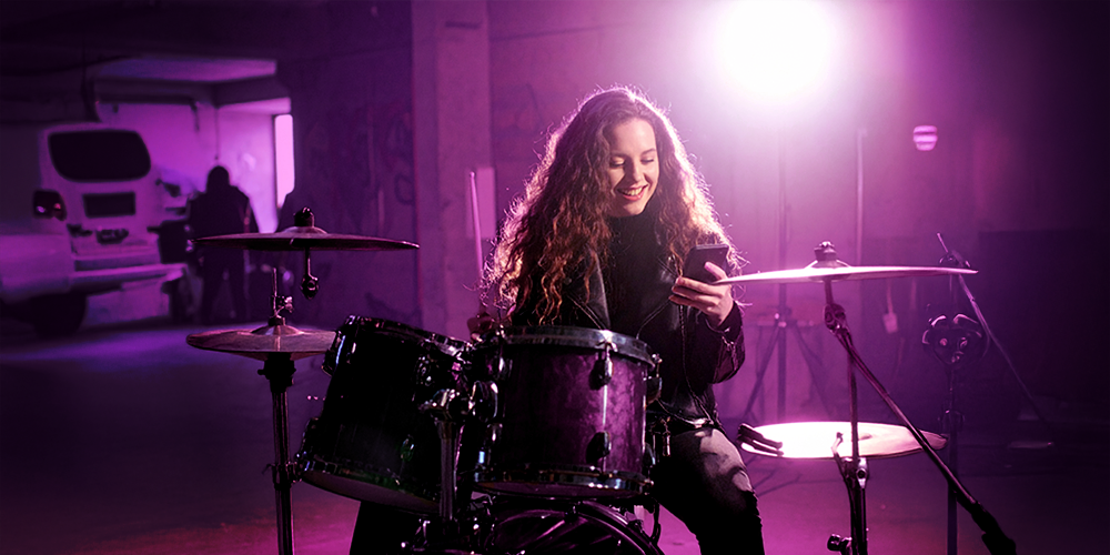 A 20-year-old woman, sitting on a rock drum set and holding her phone, excitedly playing at Playuzu.